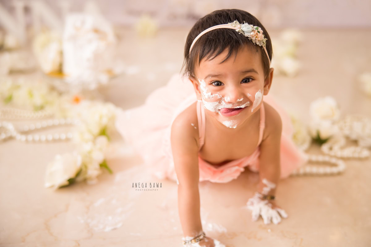 Girl crawling with pearls, white flowers, cake smash on face. First birthday photoshoot in Delhi, Gurgaon. Captured by Anega Bawa family photographer Gurgaon (Delhi NCR).