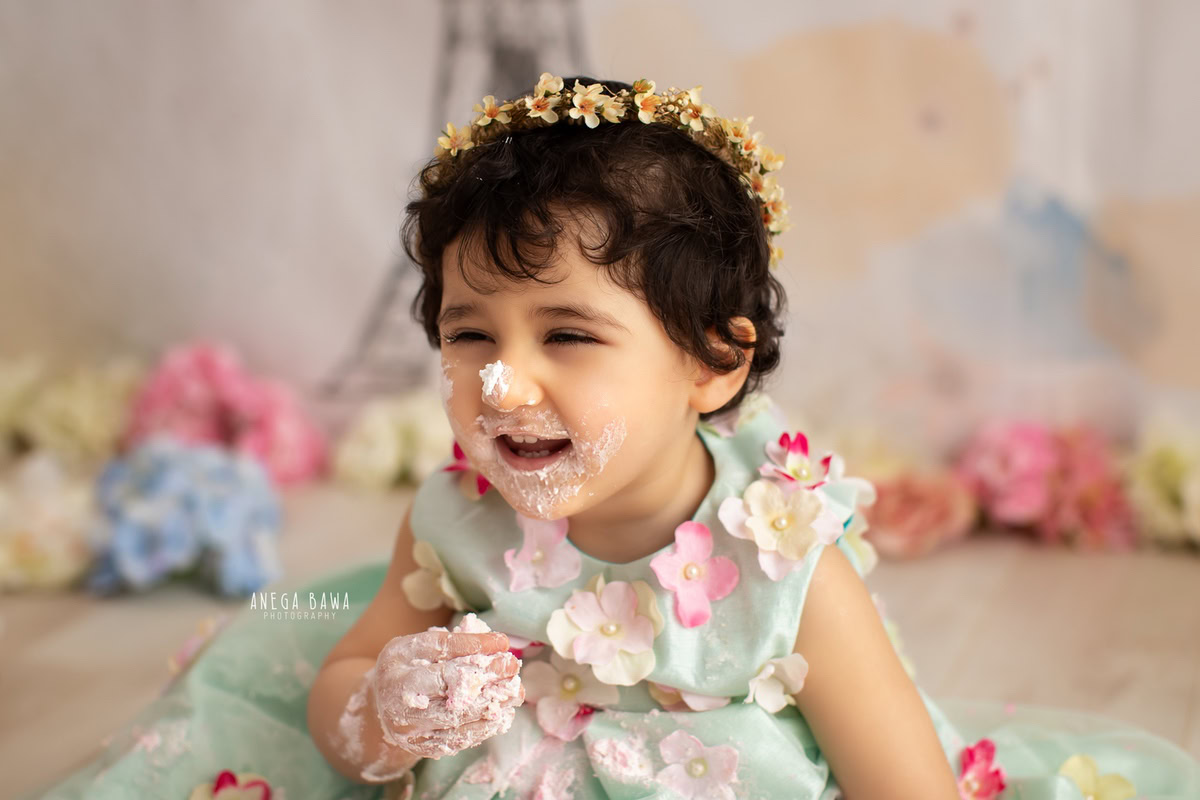 Girl with a cute smiling pose and cake on face in Cake Smash photography, surrounded by flowers on the floor, first birthday photography in Delhi, Gurgaon. Captured by Anega Bawa family photographer Gurgaon (Delhi NCR)