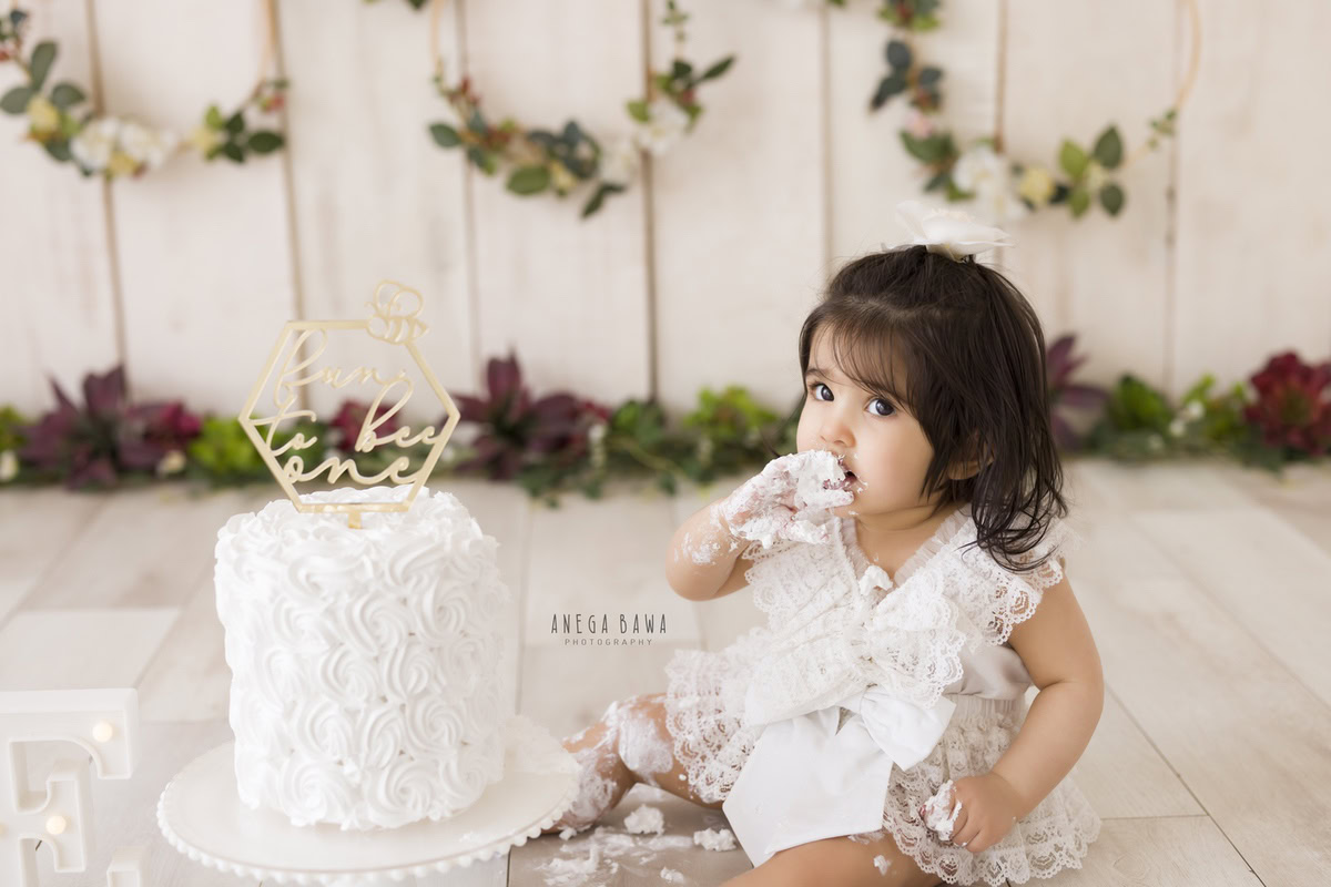 1 year old girl enjoying cake smash against a beige backdrop with a round wooden frame on the wall. Captured during a delightful first birthday photoshoot in Delhi Gurgaon by the renowned family photographer Anega Bawa.