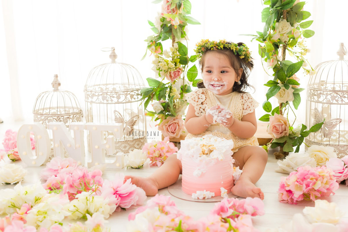 Girl enjoying a cake with white castles, flowers on the floor, and leafy fringes against a white backdrop during a Cake Smash photoshoot by well-known Anega Bawa Photography in Delhi, Gurgaon.