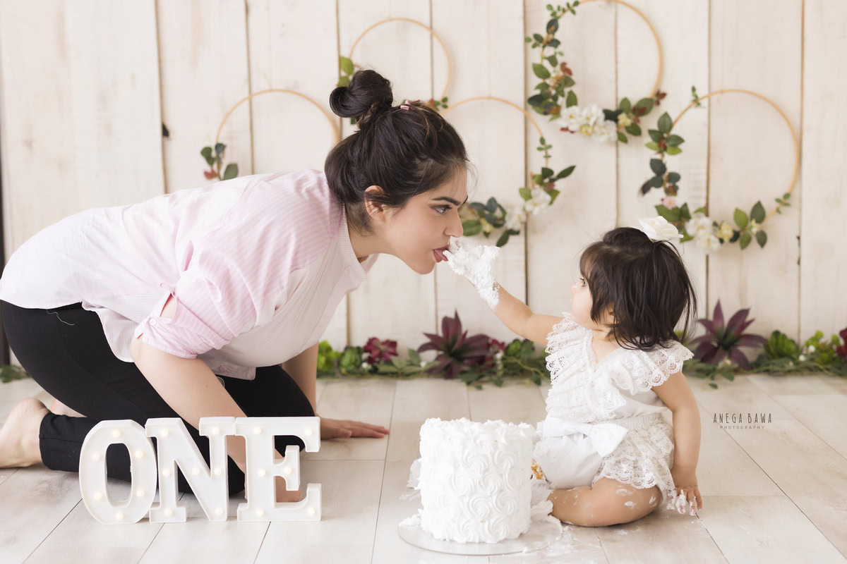 Girl enjoying a cake with her mom, set against a beige backdrop with leafy fringe on the floor during a Cake Smash photoshoot by well-known Anega Bawa Photography in Delhi, Gurgaon.