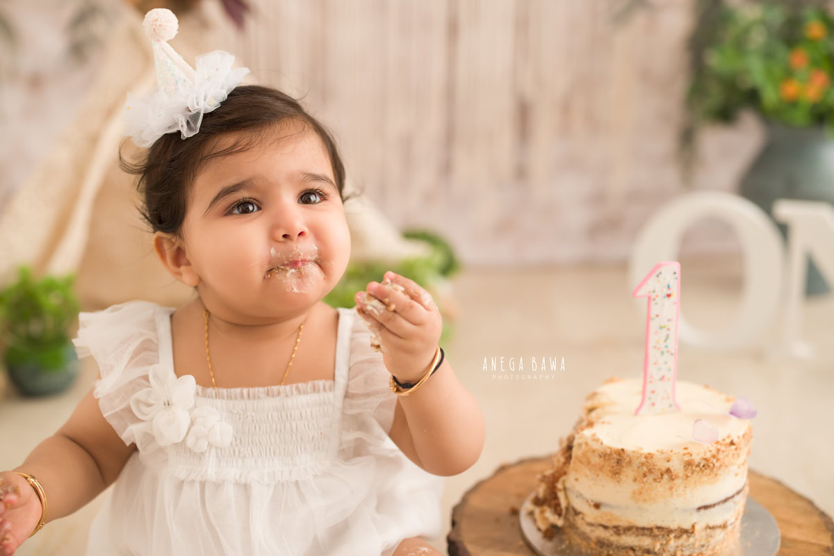 Girl in cute white dress with cake smash, hair accessory, peach and white backdrop. First birthday photoshoot in Delhi, Gurgaon. Captured by Anega Bawa family photographer Gurgaon (Delhi NCR).