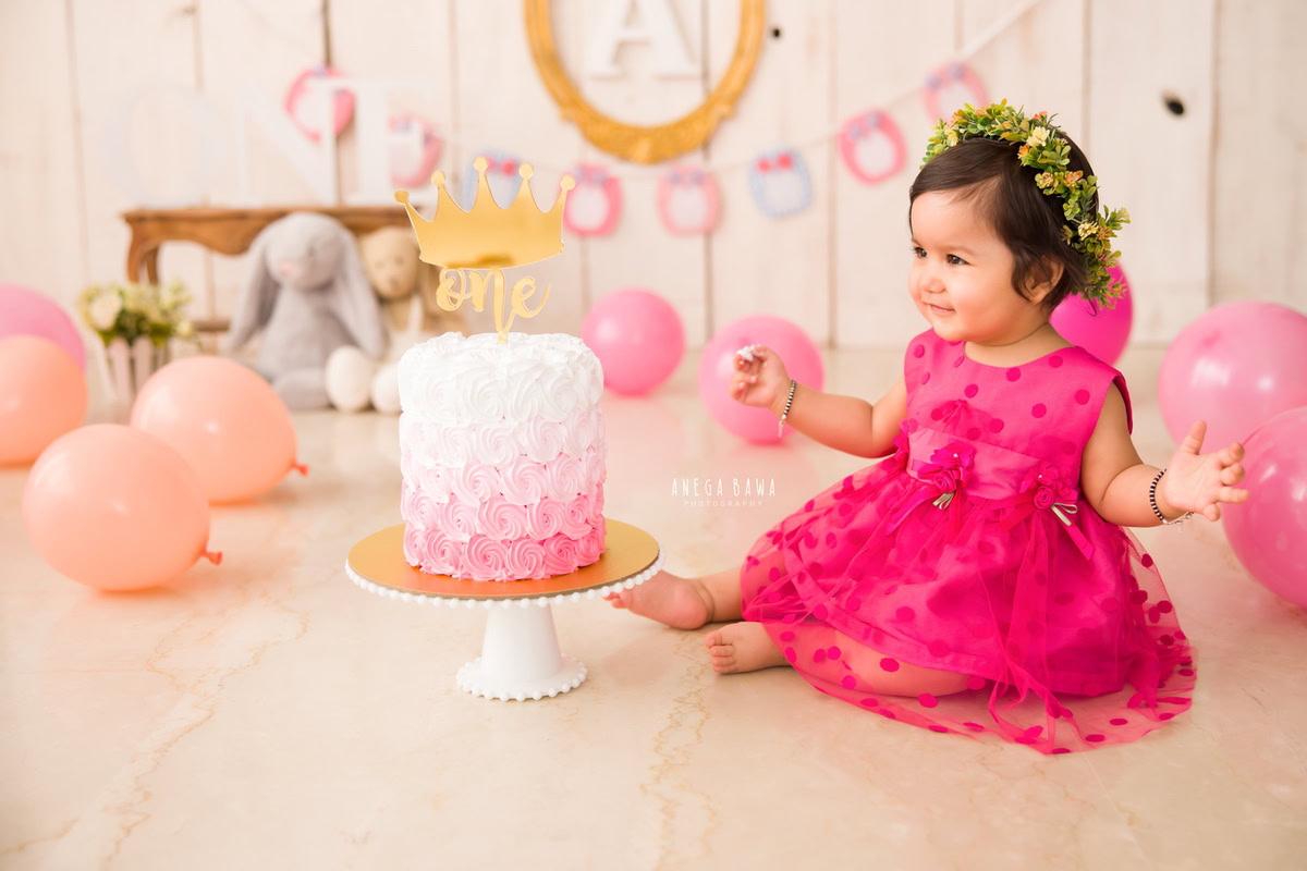Girl in pink dress with cake smash, balloons, alphabet frame on wall, beige backdrop. First birthday photoshoot in Delhi, Gurgaon. Captured by Anega Bawa family photographer Gurgaon (Delhi NCR).