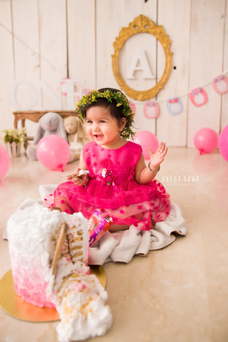 Girl in a pink dress with Cake Smash, surrounded by teddy bears, balloons, a golden alphabet frame on the wall, and a beige backdrop, first birthday photography in Delhi, Gurgaon. Captured by Anega Bawa family photographer Gurgaon (Delhi NCR)