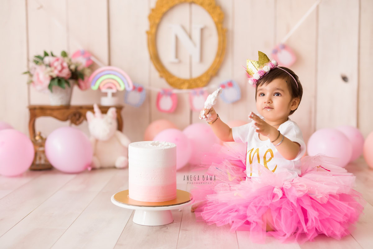 Girl sibling with cake smash on face, cute pose, leafy peach backdrop. First birthday photo shoot in Delhi, Gurgaon. Captured by Anega Bawa family photographer Gurgaon (Delhi NCR).