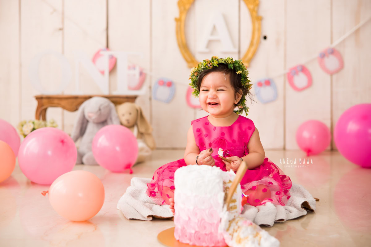 Girl in a pink dress enjoying a Cake Smash with peach and pink balloons, teddy bears, and a golden alphabet frame on the wall during a first birthday photoshoot by well-known Anega Bawa Photography in Delhi, Gurgaon.