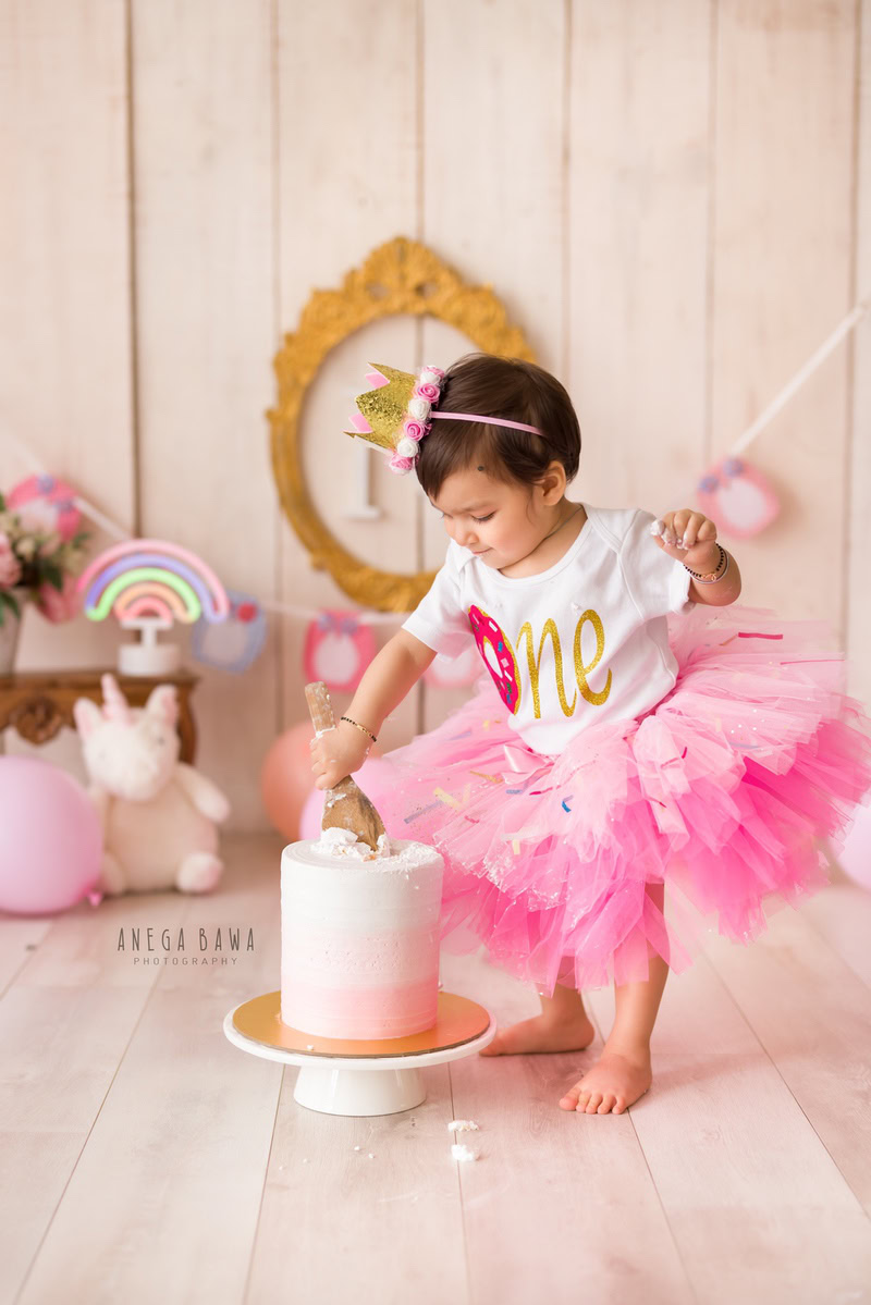 Girl playing with cake smash against a beige backdrop with a teddybear and a golden alphabet frame on the wall. Captured during a delightful first birthday photoshoot in Delhi Gurgaon by the renowned family photographer Anega Bawa.