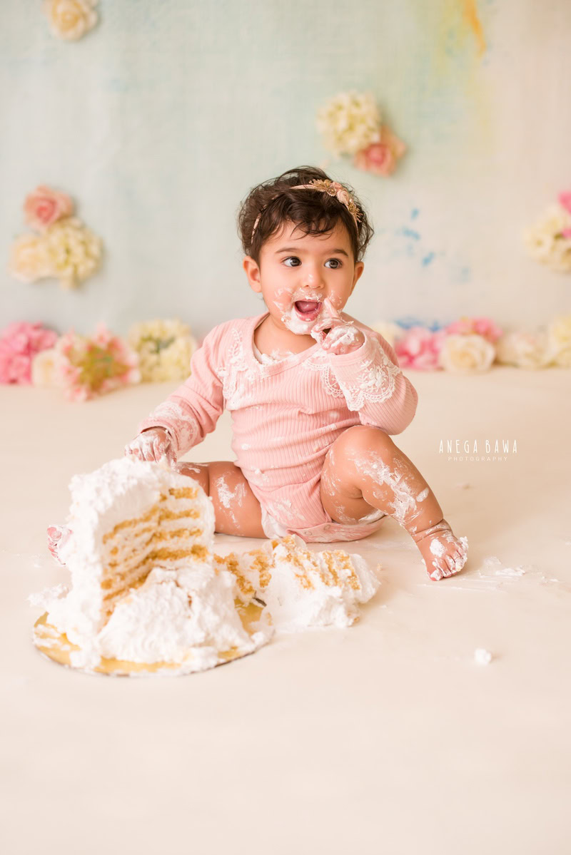 Girl playing with cake smash surrounded by multicolour flowers against a beige backdrop. Captured during a charming first birthday photoshoot in Delhi Gurgaon by the renowned family photographer Anega Bawa.