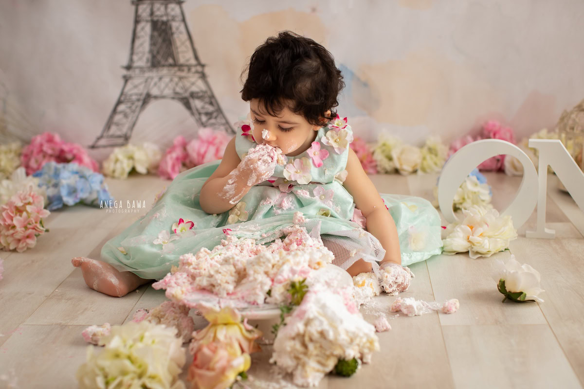 Girl playing with cake smash surrounded by multicolour flowers and an Eiffel Tower backdrop. Captured during a delightful first birthday photography session in Delhi Gurgaon by the renowned family photographer Anega Bawa.