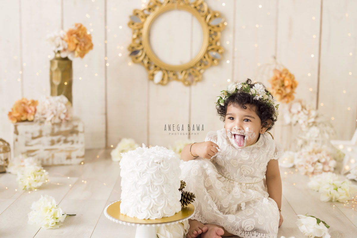 Girl playing with cake smash surrounded by orange and ivory flowers with a golden frame on the wall against a beige backdrop. Captured during a charming first birthday photoshoot in Delhi Gurgaon by the renowned family photographer Anega Bawa.
