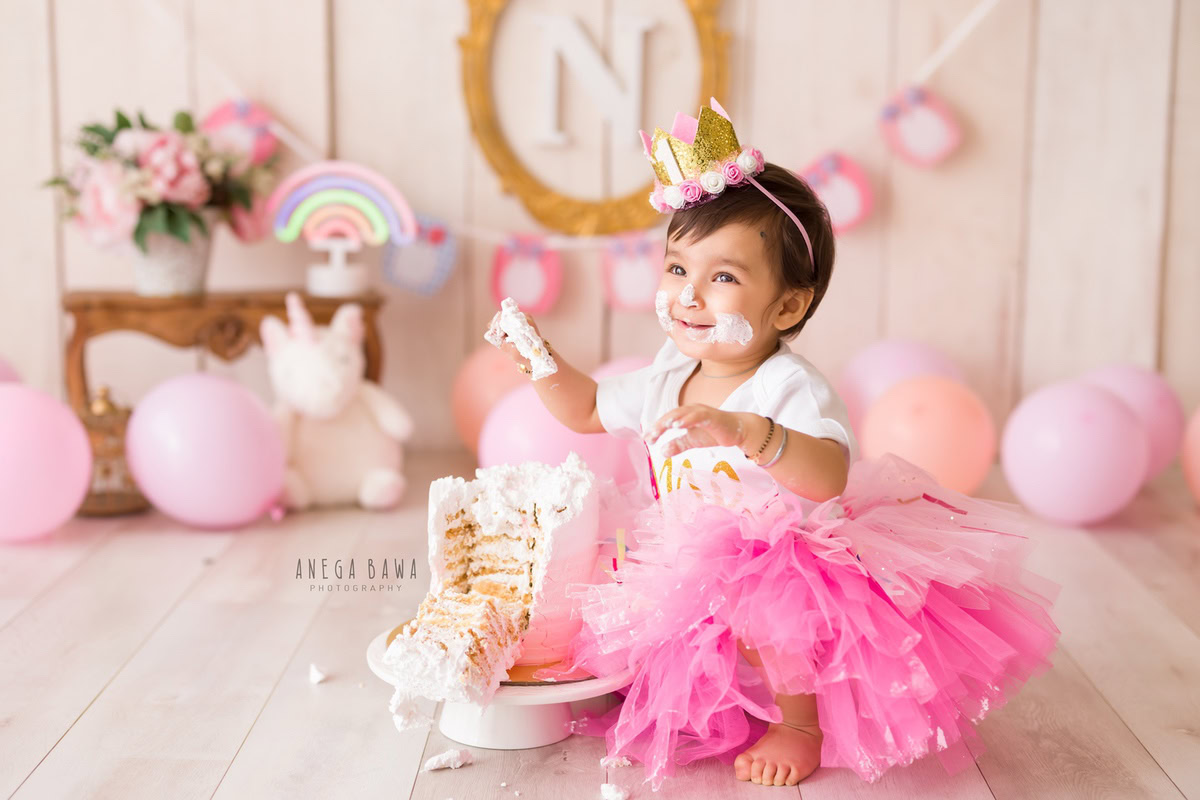 Girl playing with Cake Smash, surrounded by peach and pink balloons, a teddy bear, a beige backdrop, and an alphabet frame on the wall, Cake Smash photoshoot by Anega Bawa in Delhi, Gurgaon.