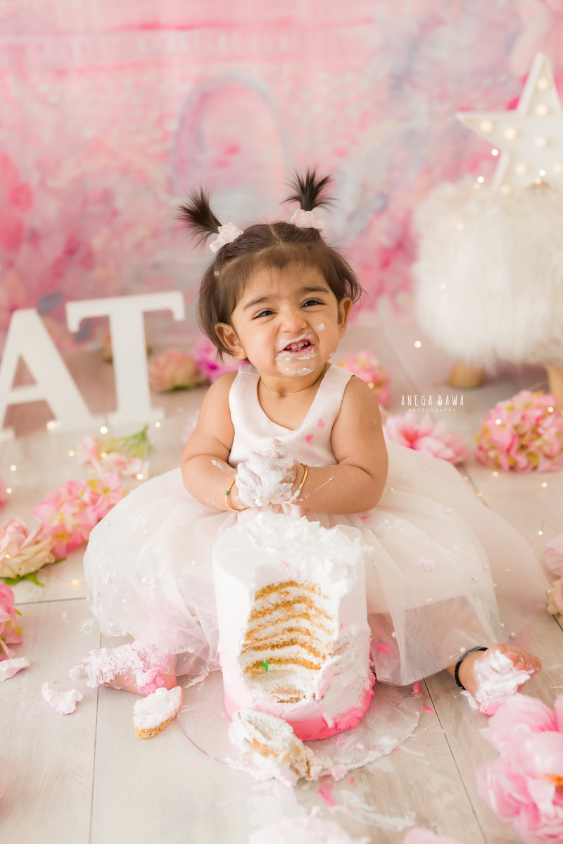 Girl enjoying Cake Smash, vibrant pink backdrop, and delicate flowers on the floor, captured in a stunning Cake Smash photoshoot by Anega Bawa in Delhi, Gurgaon.
