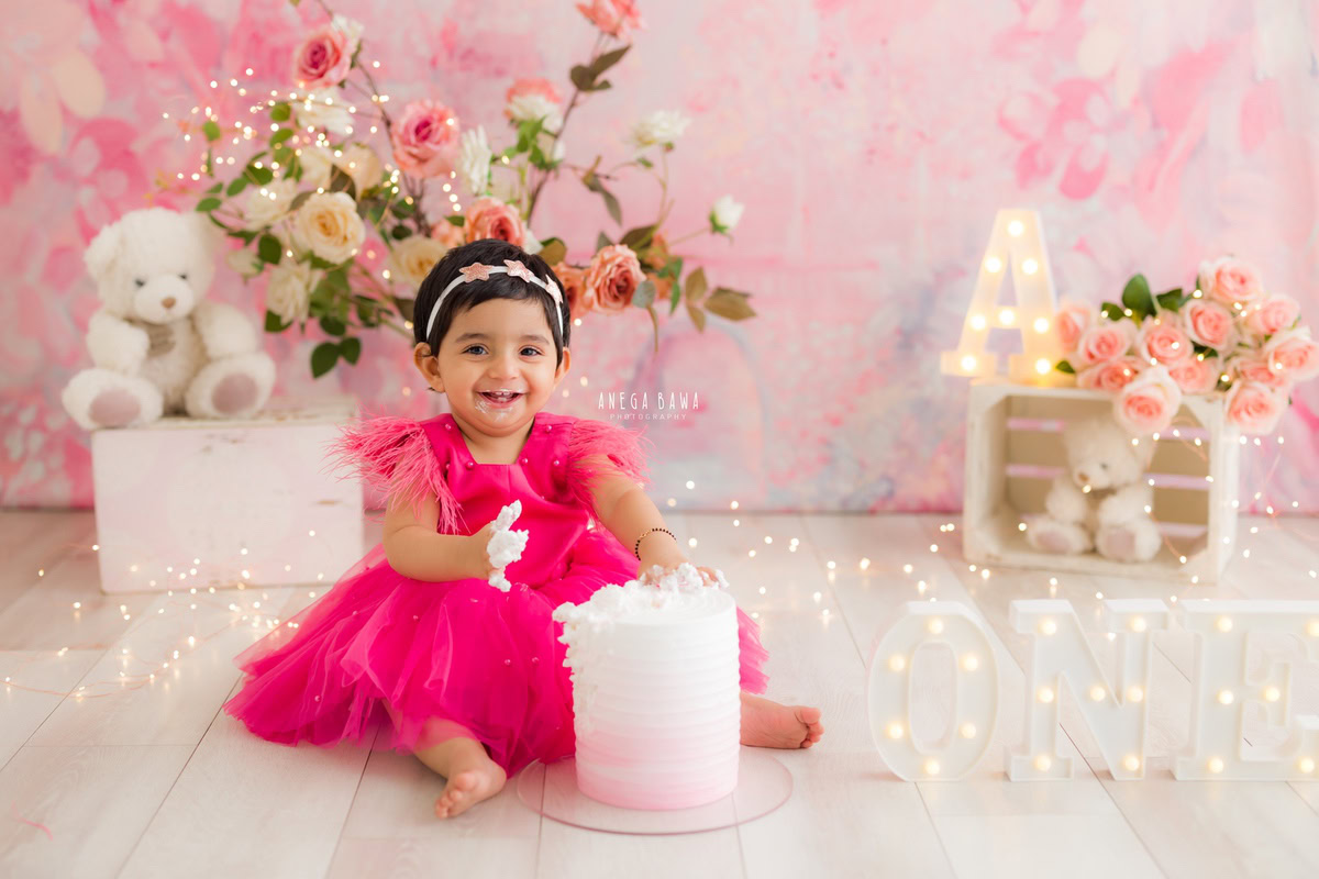 Girl playing with Cake Smash, featuring a pink backdrop, teddy bears, and a bunch of flowers, captured in a charming Cake Smash photoshoot by Anega Bawa in Delhi, Gurgaon.