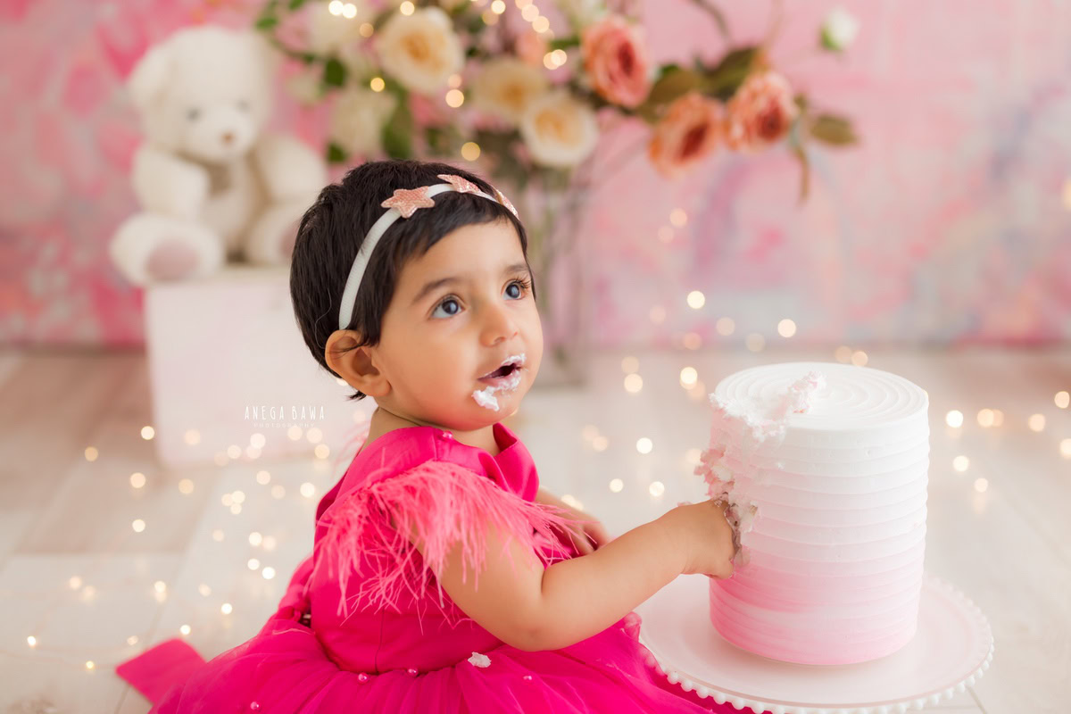 Girl playing with Cake Smash, against a pink backdrop with teddy bears and twinkling fairy lights, captured in a delightful Cake Smash photoshoot by Anega Bawa in Delhi, Gurgaon.