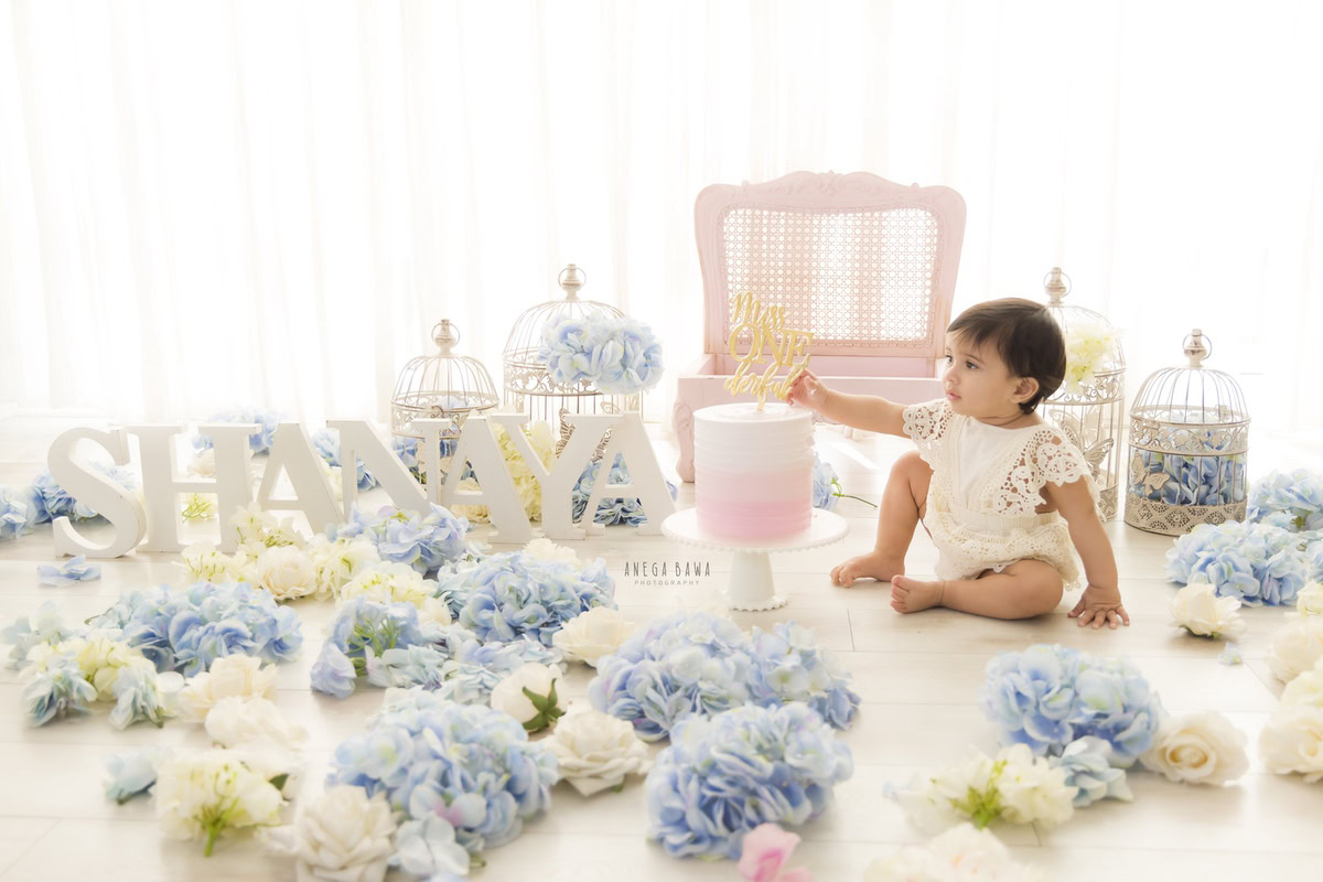 Girl playing with a cake during a Cake Smash photoshoot with a white backdrop featuring castles, blue and white flowers, and a chair by well-known Anega Bawa Photography in Delhi, Gurgaon.