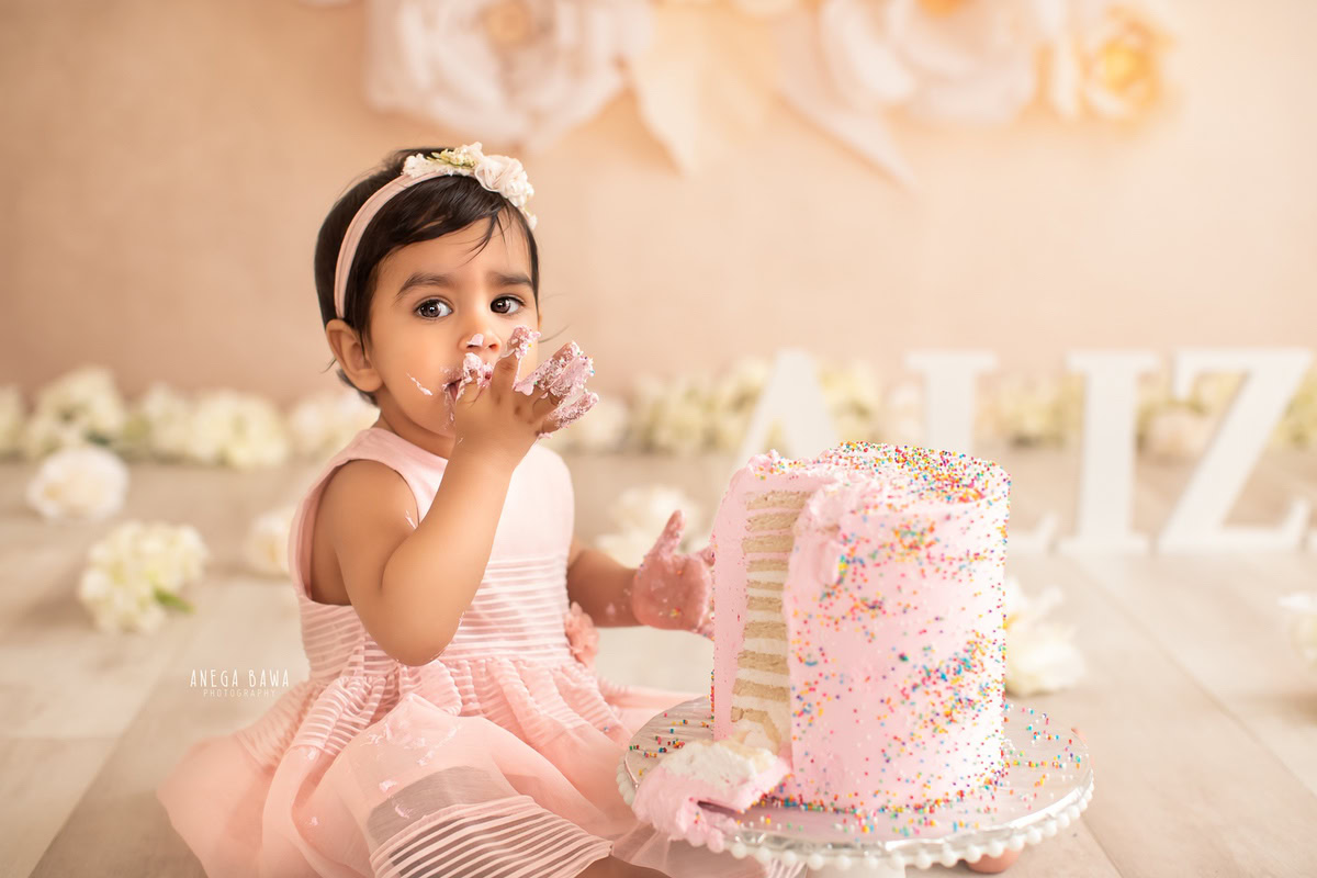 Girl playing with cake smash surrounded by white flowers and a floral beige backdrop. Captured during a delightful first birthday photography session in Delhi Gurgaon by the renowned family photographer Anega Bawa.