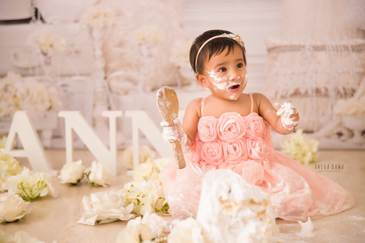 Girl playing with cake smash surrounded by white flowers and a name frame on the floor. Captured during a charming first birthday photoshoot in Delhi Gurgaon by the renowned family photographer Anega Bawa.