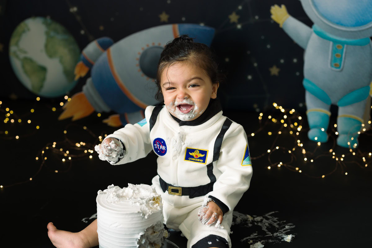 Girl posing with cake on her face and a cute smile, set against a space-themed backdrop with fairy lights during a Cake Smash photoshoot by well-known Anega Bawa Photography in Delhi, Gurgaon.