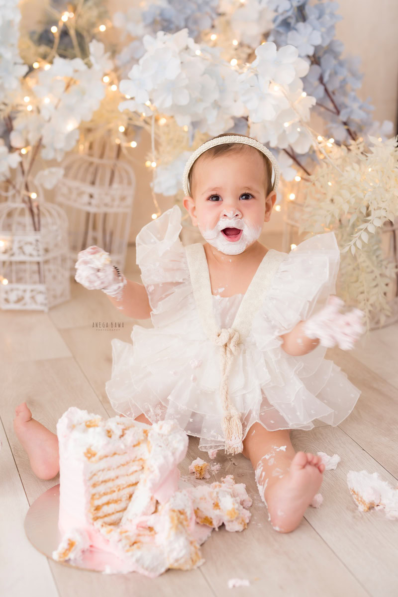 Girl posing with cake on face in Cake Smash photography, surrounded by white castles and blue and white flowers, captured in a charming first birthday photoshoot by Anega Bawa in Delhi, Gurgaon.
