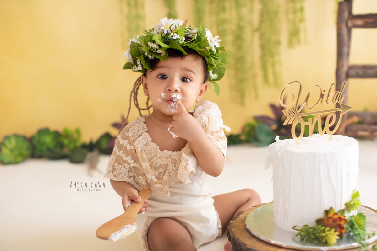 Girl posing cutely with cake smash and a tiara band against a yellow backdrop. Captured during a delightful first birthday photo shoot in Delhi Gurgaon by the renowned family photographer Anega Bawa.