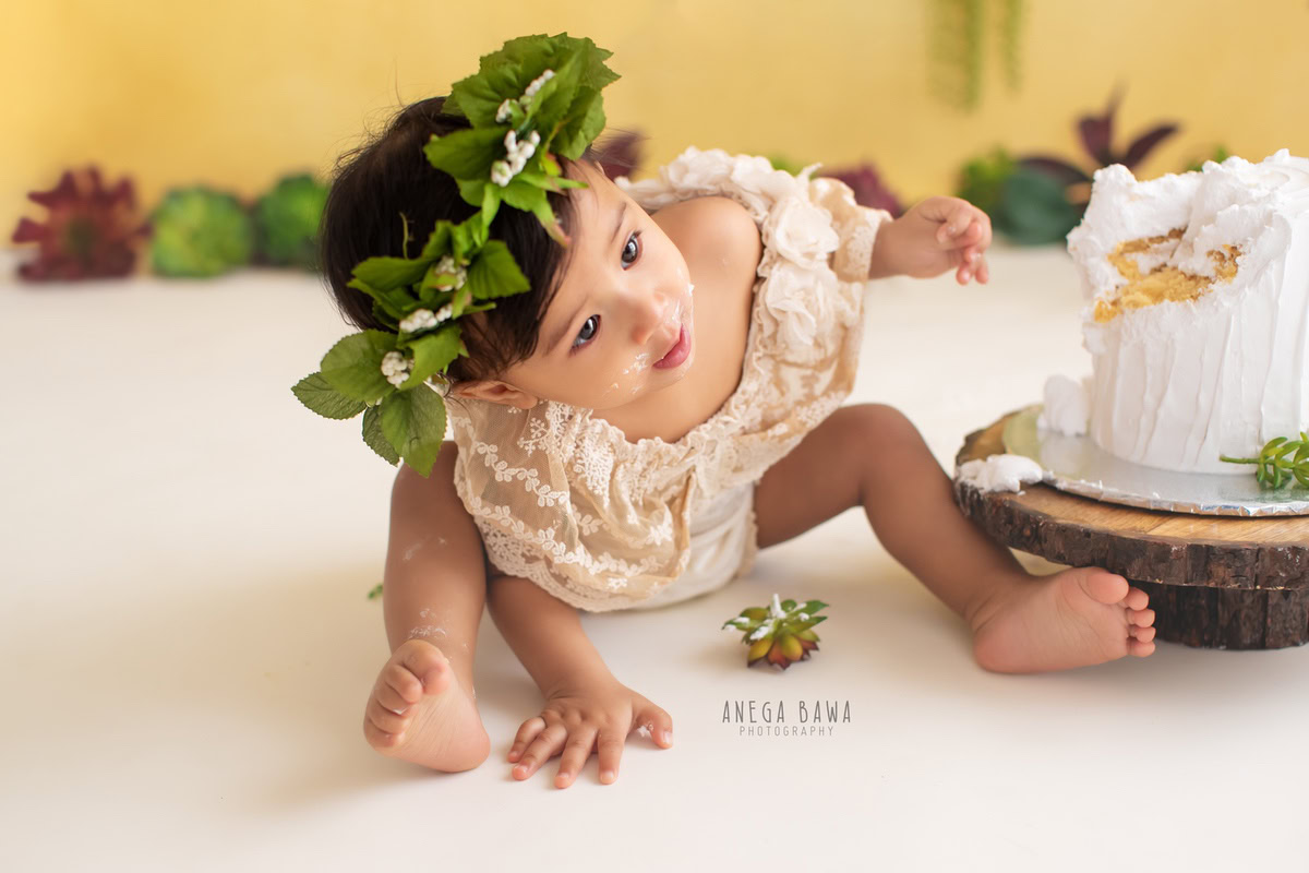 Girl posing cutely with Cake Smash, wearing a leafy tiara band, against a vibrant yellow backdrop, captured in a delightful Cake Smash photography session by Anega Bawa in Delhi, Gurgaon.