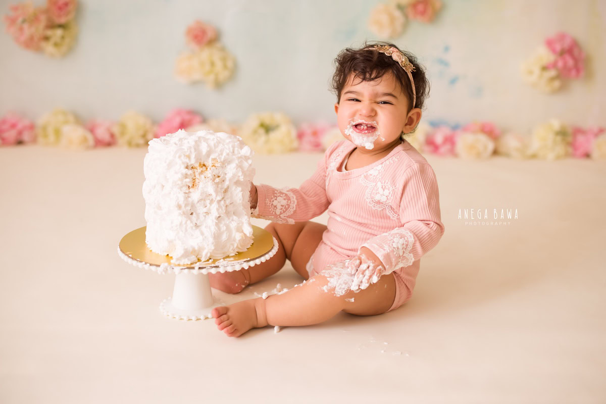Girl posing with Cake Smash, showing a cute smile, set against a white backdrop with flowers, captured in a charming Cake Smash photoshoot by Anega Bawa in Delhi, Gurgaon.
