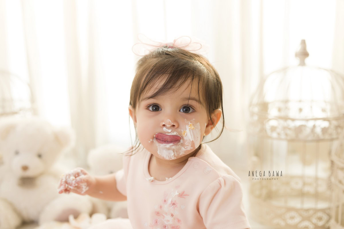 Girl posing with cake on her face, surrounded by teddy bears against a white backdrop during a Cake Smash photoshoot by well-known Anega Bawa Photography in Delhi, Gurgaon.