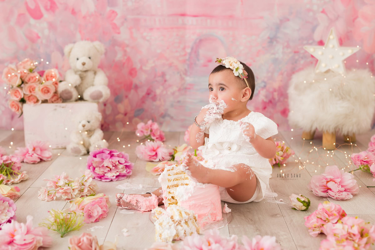 Girl posing with Cake Smash, against a pink backdrop, surrounded by teddy bears and twinkling fairy lights, captured in a delightful Cake Smash photoshoot by Anega Bawa in Delhi, Gurgaon.