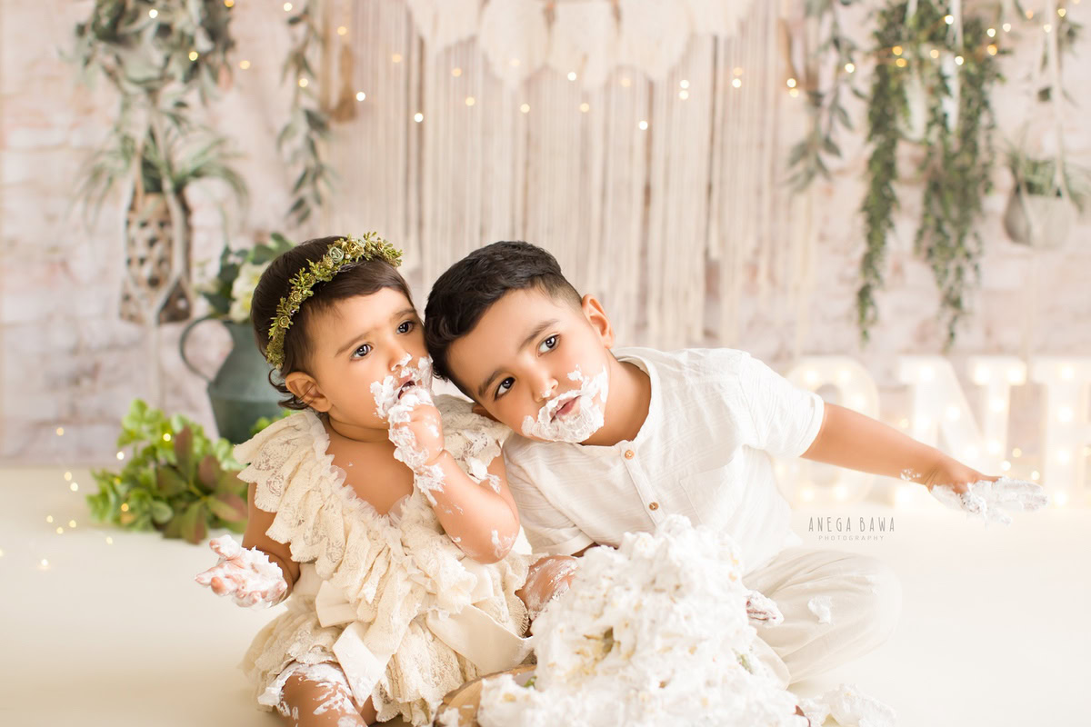 Girl sibling with cake smash on face, cute pose, leafy peach backdrop. First birthday photo shoot in Delhi, Gurgaon. Captured by Anega Bawa family photographer Gurgaon (Delhi NCR).