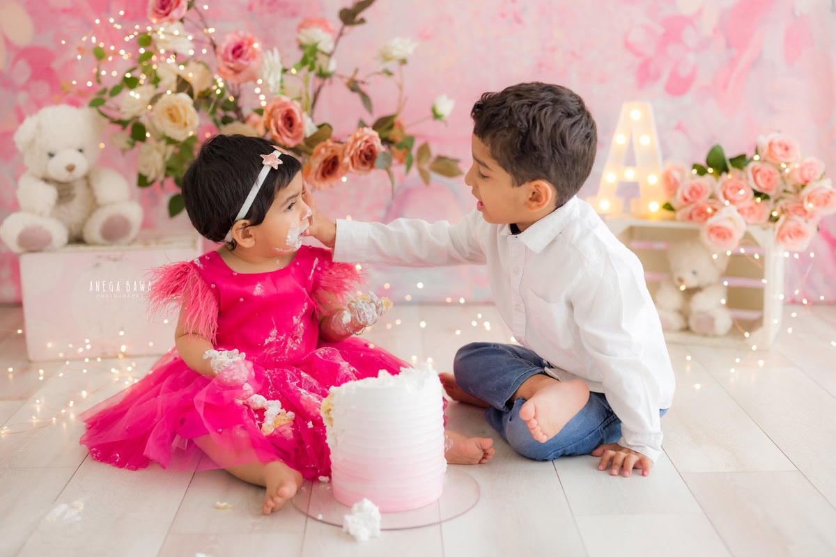Girl sibling with cake smash against a pink backdrop with fairy lights and a bunch of flowers. Captured during a delightful first birthday photo shoot in Delhi Gurgaon by the renowned family photographer Anega Bawa.
