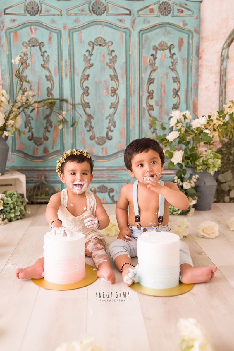 Girl sibling with green door backdrop, flower vase, two cake smashes. First birthday photo shoot in Delhi, Gurgaon. Captured by Anega Bawa family photographer Gurgaon (Delhi NCR).