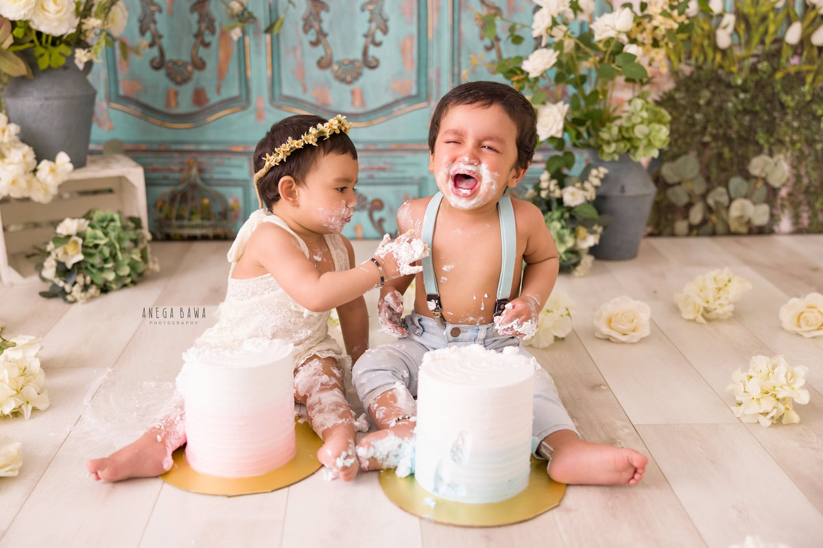 Girl sibling with two cake smashes, green door backdrop, white flowers on floor, flower vase. First birthday photo shoot in Delhi, Gurgaon. Captured by Anega Bawa family photographer Gurgaon (Delhi NCR).