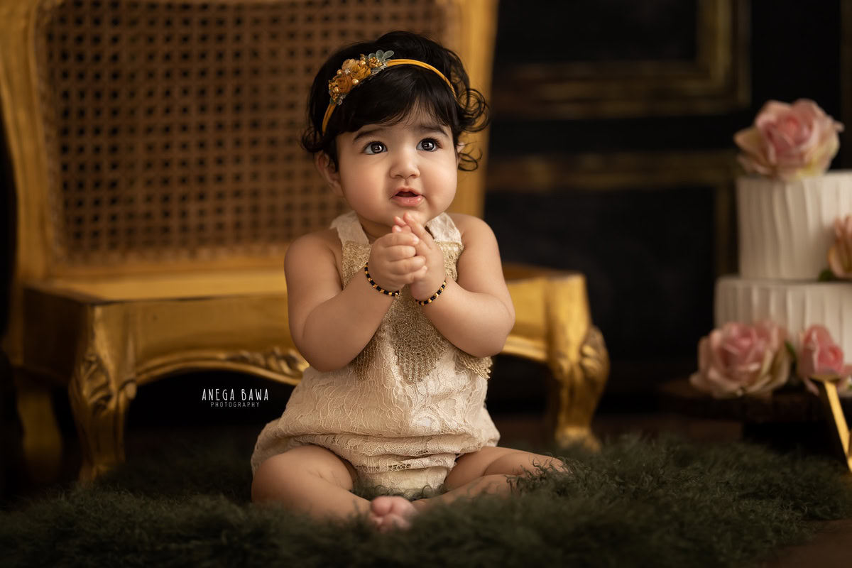 Girl sitting on a golden chair with cake smash against a brown backdrop. Captured during a charming first birthday photo shoot in Delhi Gurgaon by the renowned family photographer Anega Bawa.