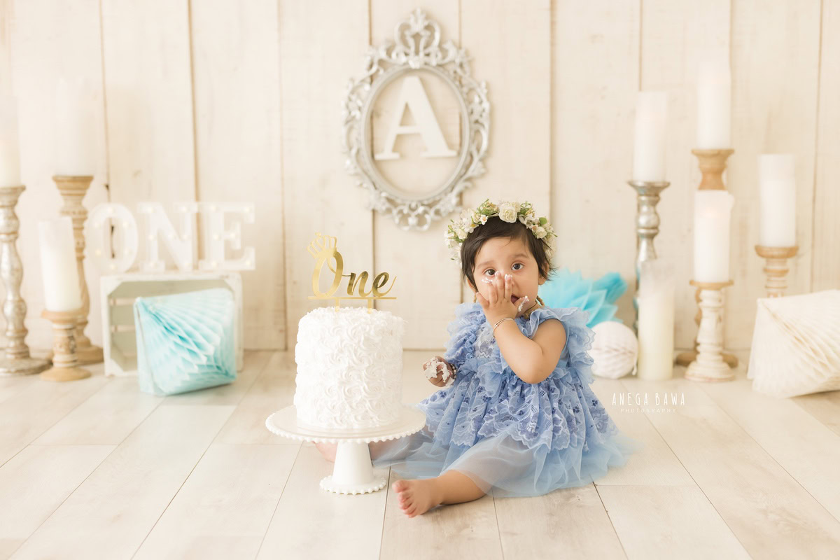 Girl sitting on the floor with a cake and a candle stand against a beige backdrop with a silver frame on the wall during a Cake Smash photoshoot by well-known Anega Bawa Photography in Delhi, Gurgaon.