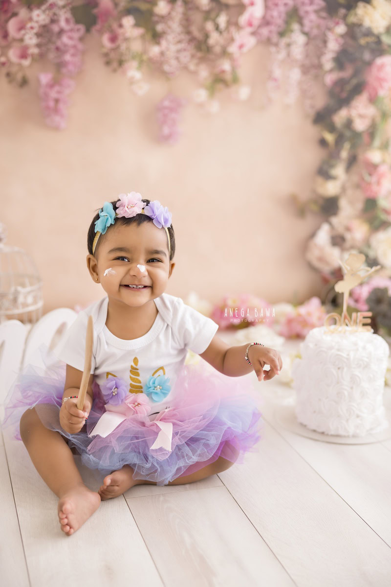 Girl sitting on the floor with Cake Smash, against a peach backdrop with pink floral fringe on the wall, captured in a charming Cake Smash photography session by Anega Bawa in Delhi, Gurgaon.