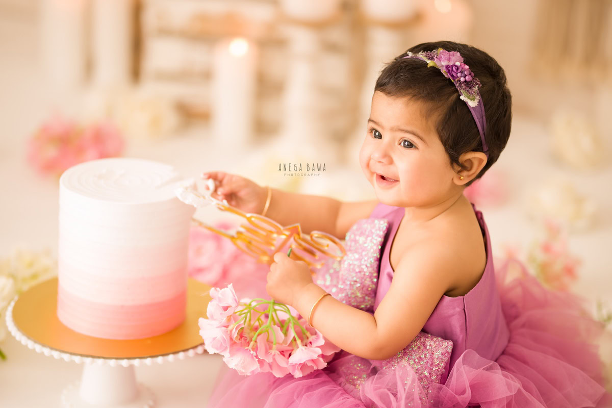 Girl sitting on floor with cake smash, holding flowers in hand, candles, beige backdrop. First birthday photoshoot in Delhi, Gurgaon. Captured by Anega Bawa family photographer Gurgaon (Delhi NCR).