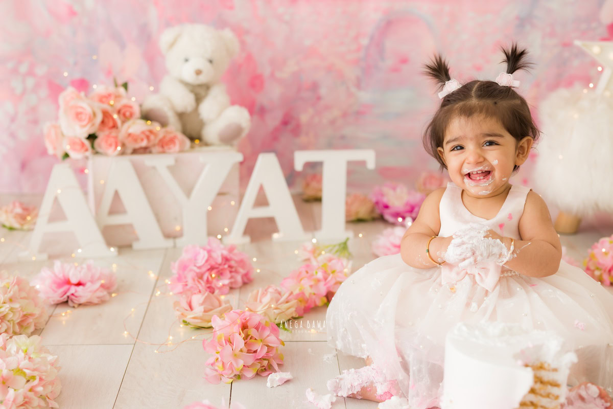 Girl sitting on the floor with Cake Smash, set against a pink backdrop, surrounded by teddy bears and a name frame on the floor, captured in a delightful Cake Smash photoshoot by Anega Bawa in Delhi, Gurgaon.
