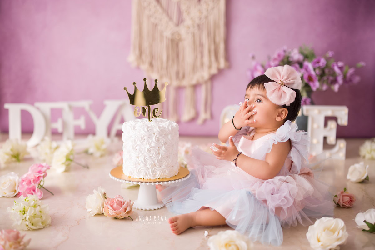 Girl sitting on floor with name frame, lavender backdrop, cake smash, flowers on floor. First birthday photography in Delhi, Gurgaon. Captured by Anega Bawa family photographer Gurgaon (Delhi NCR).