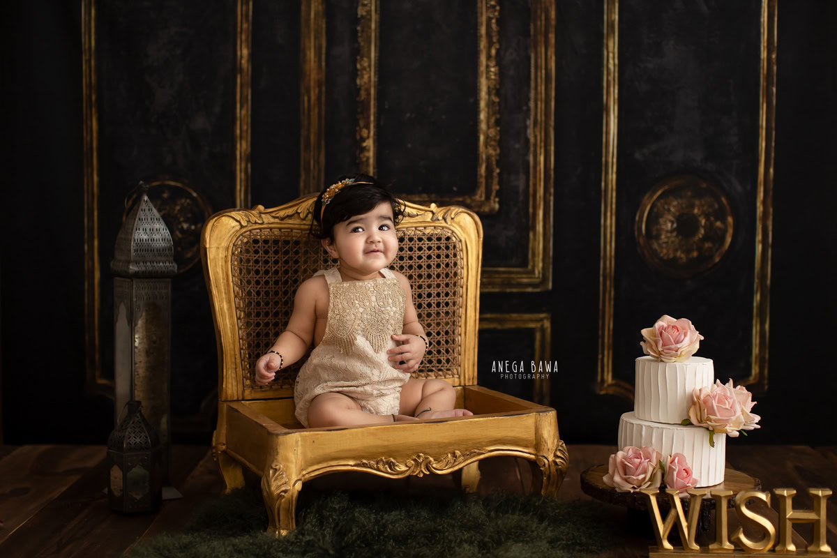 Girl sitting on golden chair with cake smash, brown backdrop. First birthday photo shoot in Delhi, Gurgaon. Captured by Anega Bawa family photographer Gurgaon (Delhi NCR).