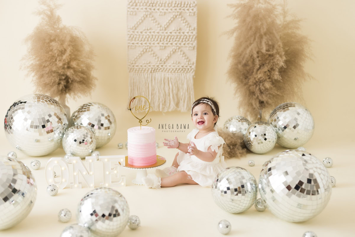 Girl sitting with a cake, surrounded by disco balls and bushes against a beige backdrop during a Cake Smash photoshoot by well-known Anega Bawa Photography in Delhi, Gurgaon.