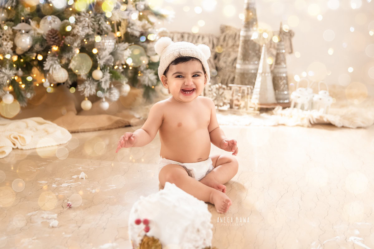 Girl sitting with cake smash against an Xmas-themed backdrop with fairy lights. Captured during a festive first birthday photo shoot in Delhi Gurgaon by the renowned family photographer Anega Bawa.
