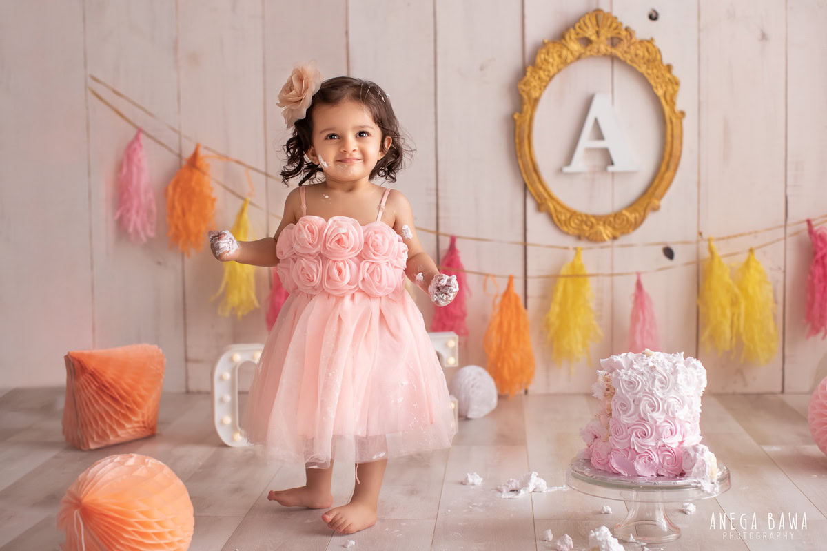 Girl standing with Cake Smash, featuring an alphabet frame and colorful fringe on the wall, complemented by pompom decorations, captured in a vibrant Cake Smash photoshoot by Anega Bawa in Delhi, Gurgaon.