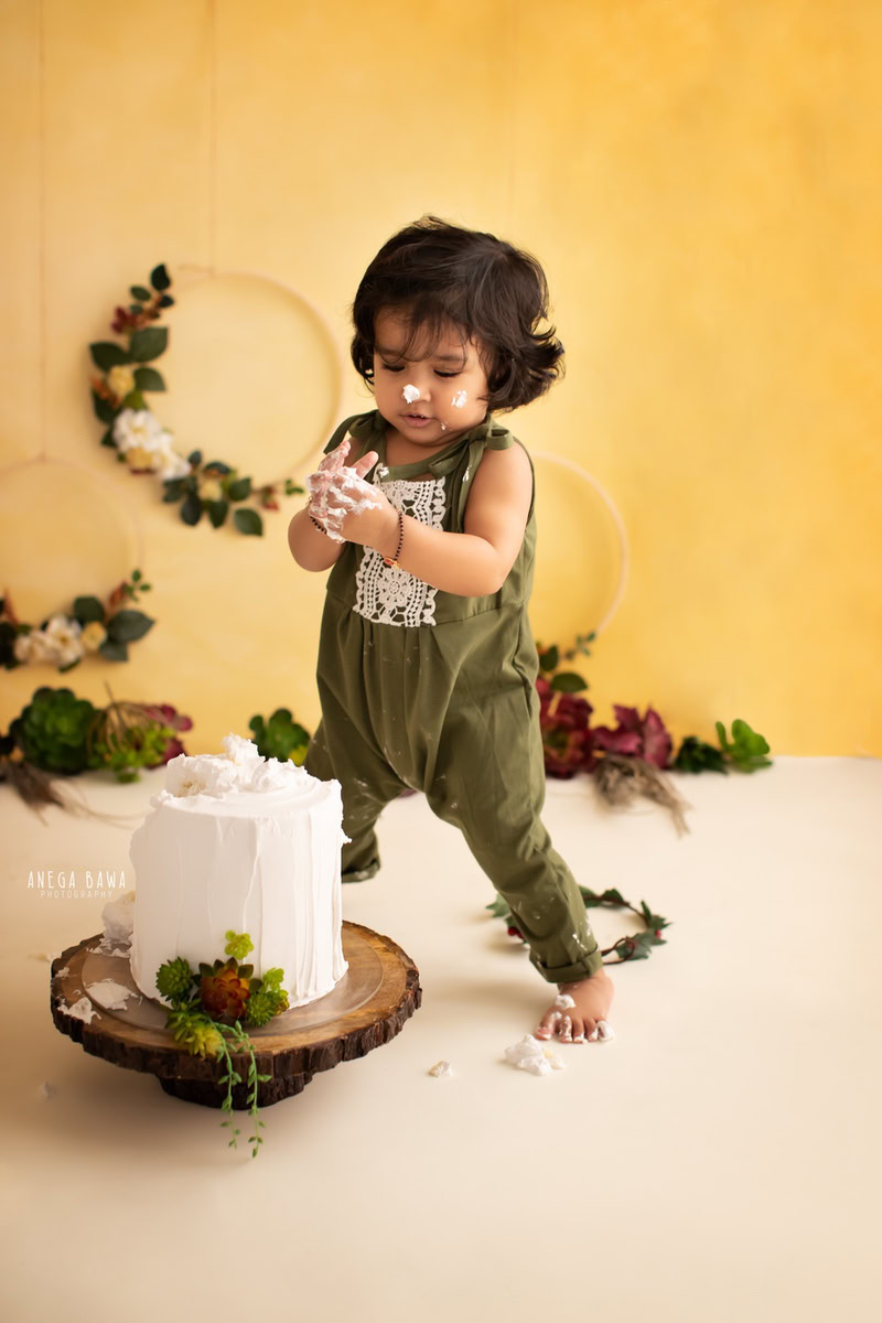 Girl standing with cake smash against a yellow backdrop featuring a floral round frame on the wall. Captured during a vibrant first birthday photoshoot in Delhi Gurgaon by the renowned family photographer Anega Bawa.