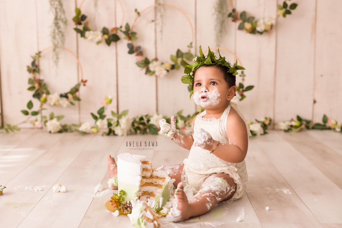 Girl with tiara band and cake smash, leafy wooden frames on wall, beige backdrop. First birthday photoshoot in Delhi, Gurgaon. Captured by Anega Bawa family photographer Gurgaon (Delhi NCR).
