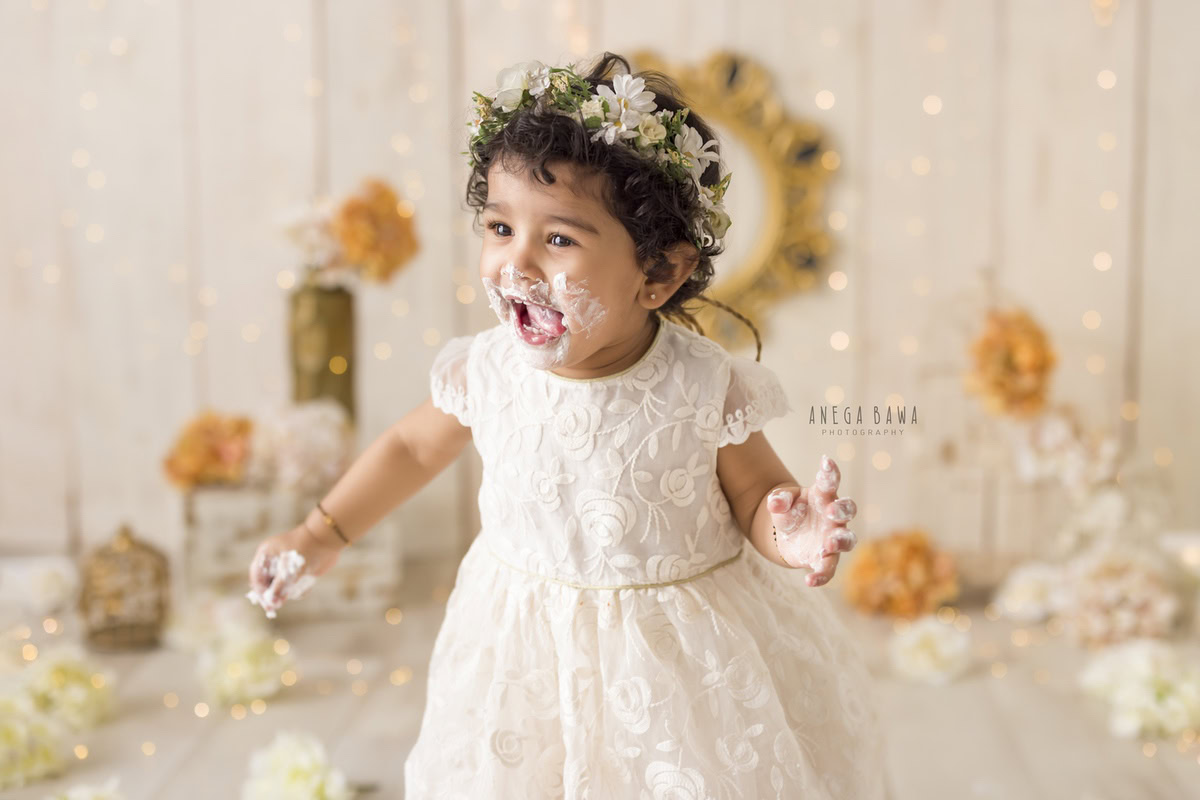 Girl with a tiara band and a cute smiling pose, with cake on her face, surrounded by orange and ivory flowers and fairy lights against a beige backdrop during a Cake Smash photoshoot by well-known Anega Bawa Photography in Delhi, Gurgaon.