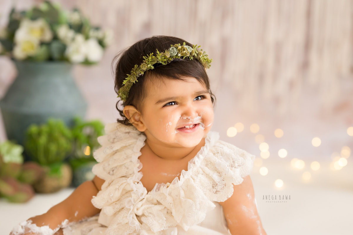 Girl with a tiara band and cake on face, surrounded by a green vase, white flowers, and fairy lights, displaying a cute smile, captured in a charming Cake Smash photography session by Anega Bawa in Delhi, Gurgaon.