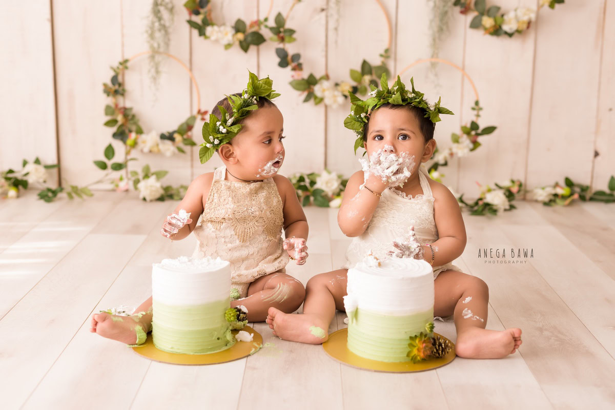 Twin girls with cake smash on their faces against a beige backdrop featuring a leafy wooden circular frame on the wall. Captured during a delightful first birthday photography session in Delhi Gurgaon by the renowned family photographer Anega Bawa.
