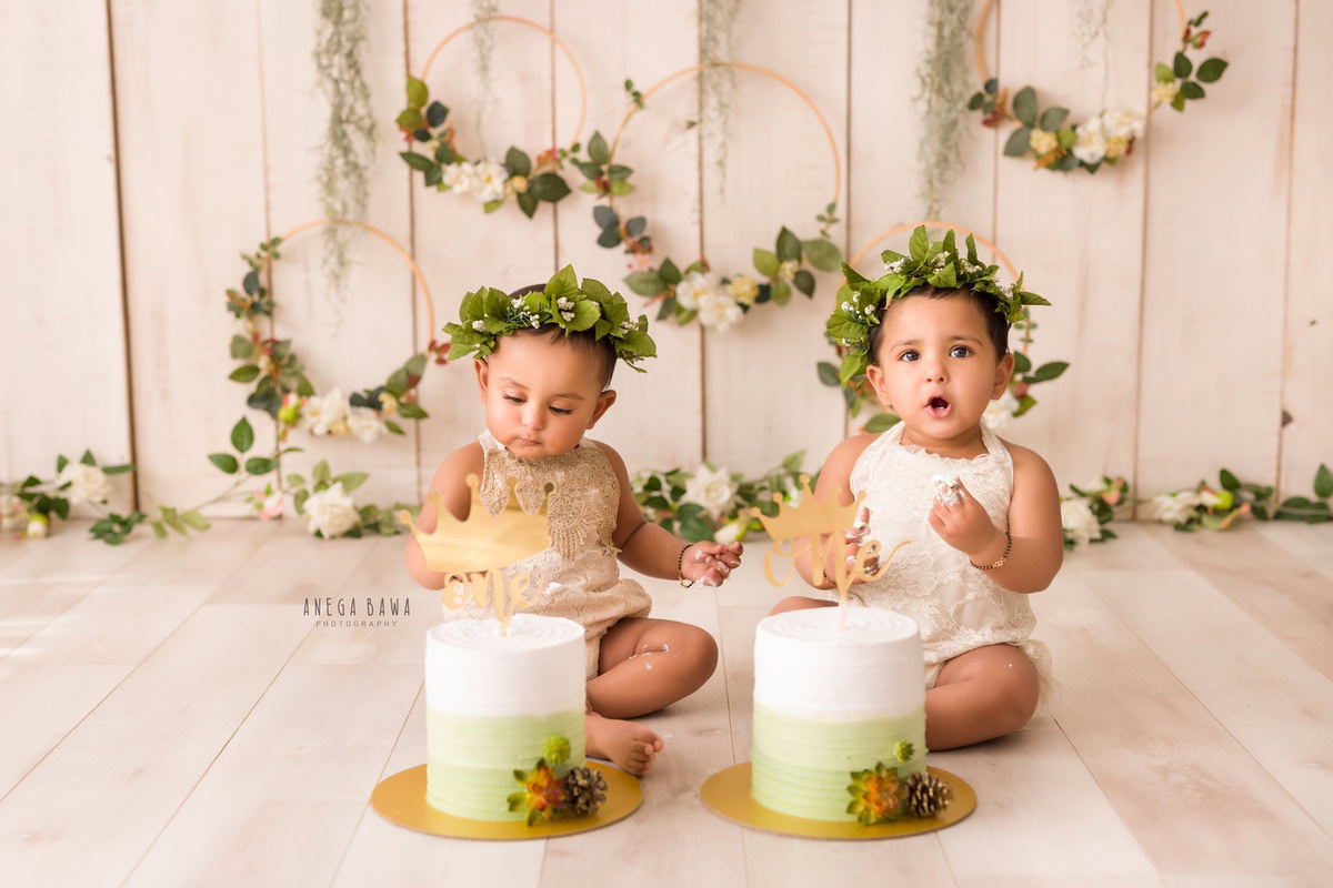 Girl twins with two cake smashes, beige backdrop, leafy circular frame on wall. First birthday photography in Delhi, Gurgaon. Captured by Anega Bawa family photographer Gurgaon (Delhi NCR).