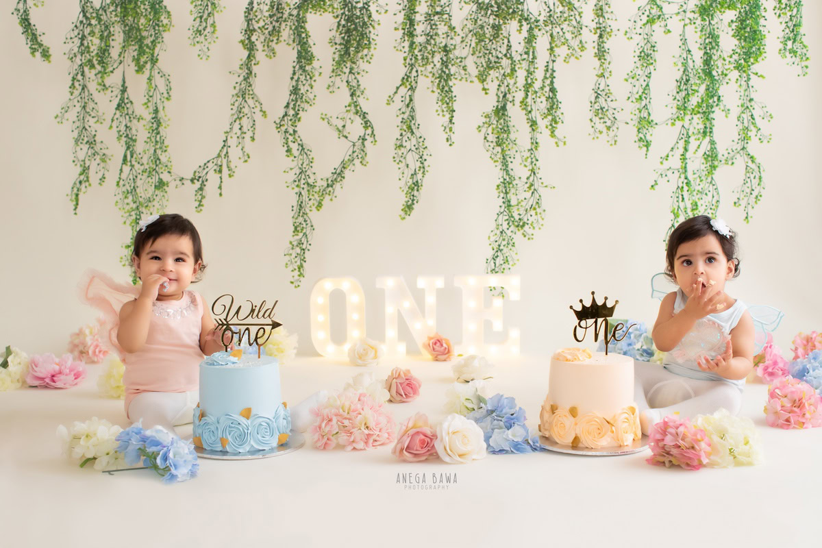 Twin girls with two cake smashes surrounded by flowers on the floor and a leafy fringe on the wall against a white backdrop. Captured during a charming first birthday photoshoot in Delhi Gurgaon by the renowned family photographer Anega Bawa.