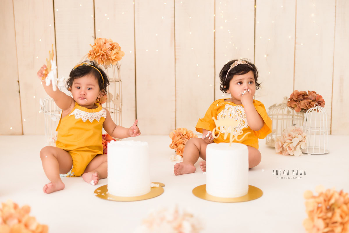 Girl twins with beige backdrop, orange and brown flowers, white castle, two cake smashes. First birthday photoshoot in Delhi, Gurgaon. Captured by Anega Bawa family photographer Gurgaon (Delhi NCR).